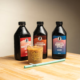 Three bottles of Rook Coffee cold brew, one 'New Orleans,' one 'Vanilla,' one 'Gingerbread' next to a glass with 'Rook Coffee Est. 2010' in gold script, a green silicone straw sticking out